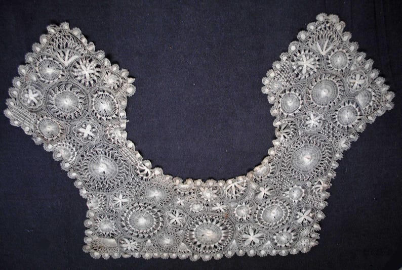May include: A delicate, white lace collar with an intricate floral and circular pattern. The collar is U-shaped, with detailed openwork and scalloped edges. The lace is set against a dark background.