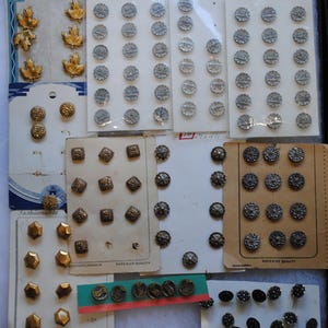 May include: A collection of vintage buttons in various shapes, sizes, and colors. The assortment includes gold leaf-shaped buttons, round silver buttons, square textured buttons, and faceted gold buttons. Some buttons are attached to their original cards.