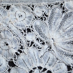 May include: Close-up of white lace textile with floral and leaf patterns. The lace features detailed openwork designs, including circular and fan-shaped motifs. The delicate fabric is set against a dark background, highlighting the intricate details of the lace.