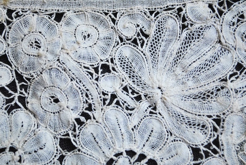 May include: Close-up of white lace textile with floral and leaf patterns. The lace features detailed openwork designs, including circular and fan-shaped motifs. The delicate fabric is set against a dark background, highlighting the intricate details of the lace.