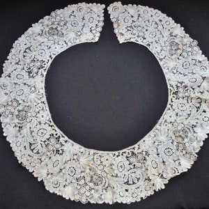 May include: A delicate, off-white lace collar with floral and leaf patterns. The collar is open in the front and features intricate detailing throughout. The lace is likely vintage or antique, with a soft, textured appearance. The collar is set against a dark background.