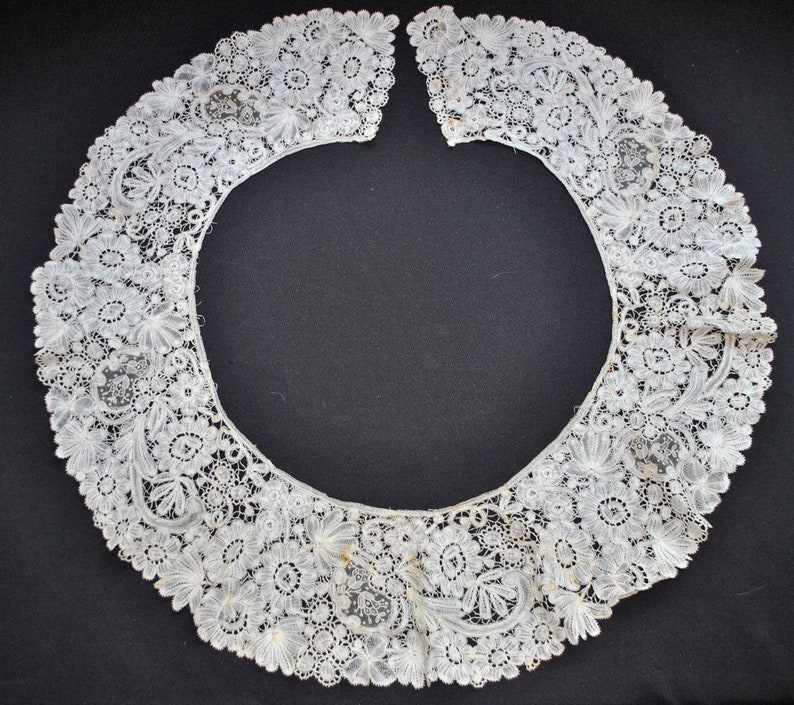 May include: A delicate, off-white lace collar with floral and leaf patterns. The collar is open in the front and features intricate detailing throughout. The lace is likely vintage or antique, with a soft, textured appearance. The collar is set against a dark background.