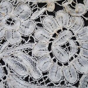 May include: Close-up of white lace fabric with a floral pattern. The lace features detailed, openwork designs of flowers and leaves, creating a delicate and intricate texture. The background is a dark, contrasting color.