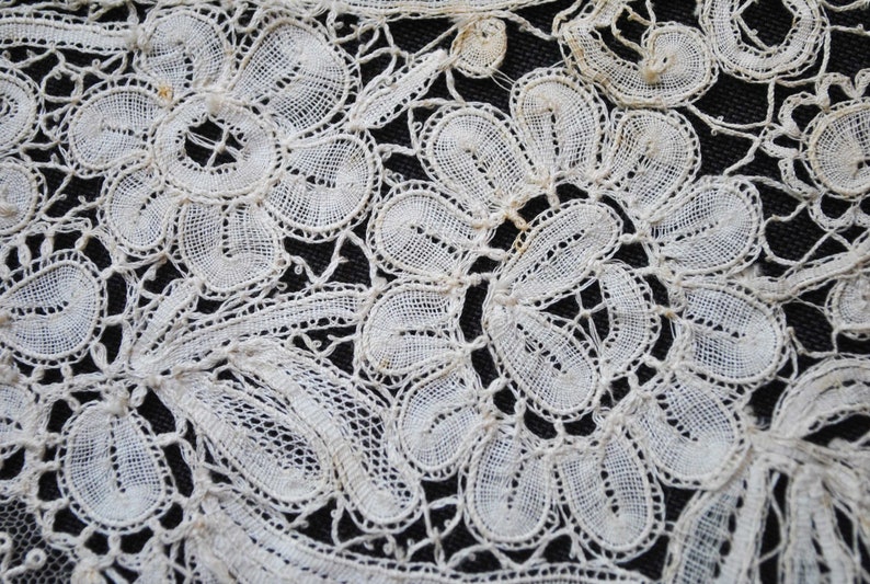 May include: Close-up of white lace fabric with a floral pattern. The lace features detailed, openwork designs of flowers and leaves, creating a delicate and intricate texture. The background is a dark, contrasting color.