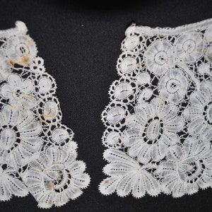 May include: Close-up of two sections of a white lace collar. The lace features a floral design with detailed patterns and scalloped edges. The delicate fabric is set against a dark background, highlighting the intricate details.
