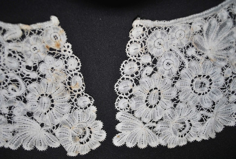 May include: Close-up of two sections of a white lace collar. The lace features a floral design with detailed patterns and scalloped edges. The delicate fabric is set against a dark background, highlighting the intricate details.