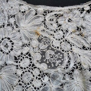 May include: Close-up of a white lace textile with a floral pattern. The lace features detailed designs of flowers and leaves, with a delicate, openwork construction. The intricate details are visible against a dark background.