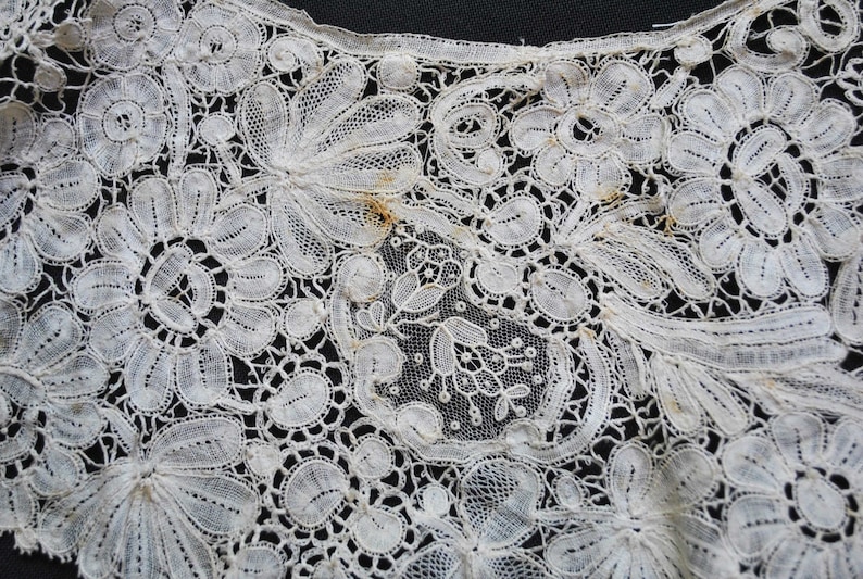 May include: Close-up of a white lace textile with a floral pattern. The lace features detailed designs of flowers and leaves, with a delicate, openwork construction. The intricate details are visible against a dark background.