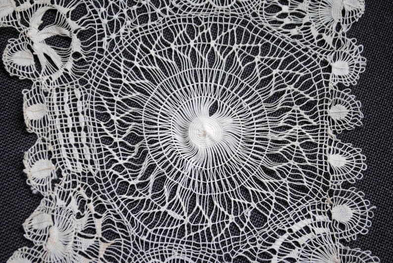 May include: Close-up of a white, intricately designed lace doily. The delicate textile features a central circular pattern with radiating lines and floral-like details around the edges. The lace is set against a dark background.