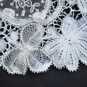 May include: Close-up of white lace with floral and circular patterns. The delicate lace features intricate openwork designs and scalloped edges, creating a textured and detailed appearance. The lace is set against a dark background.