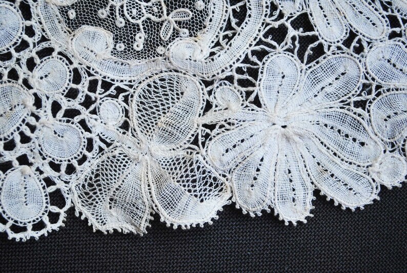 May include: Close-up of white lace with floral and circular patterns. The delicate lace features intricate openwork designs and scalloped edges, creating a textured and detailed appearance. The lace is set against a dark background.