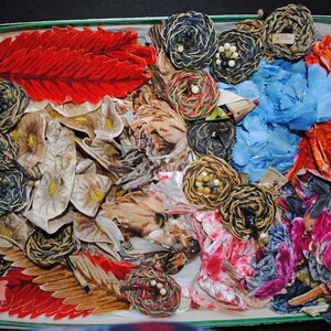 Pode incluir: Uma caixa cheia de materiais de artesanato vintage variados. A caixa contém folhas de tecido vermelhas, castanhas e bege, flores de tecido azuis e laranja e enfeites circulares tecidos com contas. A caixa tem uma borda verde.