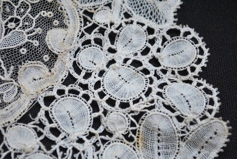 May include: Close-up of antique white lace with intricate floral and circular patterns. The delicate textile features detailed openwork and a scalloped edge, showcasing the craftsmanship of vintage lace.