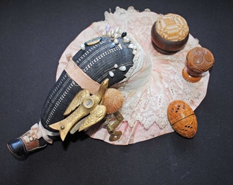 May include: An antique sewing kit with a black and white pin cushion, adorned with pins and pearls. The kit includes a brass angel-shaped needle holder, a wooden pincushion, and a carved wooden egg-shaped container, all set on a pink lace-trimmed fabric.