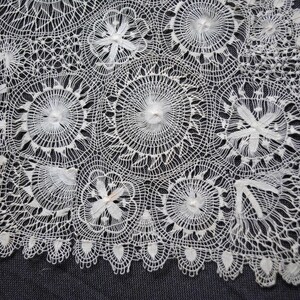 May include: A white, handmade lace doily with a detailed floral pattern. The doily features circular and floral designs, with a scalloped edge. The delicate lace is set against a dark gray background.
