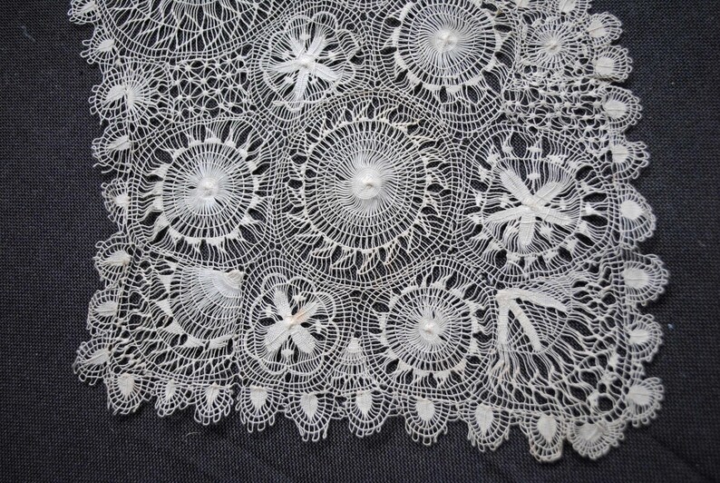 May include: A white, handmade lace doily with a detailed floral pattern. The doily features circular and floral designs, with a scalloped edge. The delicate lace is set against a dark gray background.