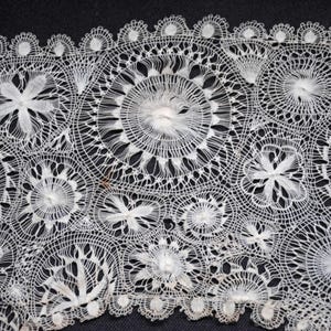 May include: Close-up of white lace fabric with intricate floral and circular patterns. The lace features a scalloped edge and detailed openwork designs, creating a delicate and textured appearance. The background is dark, highlighting the lace's details.