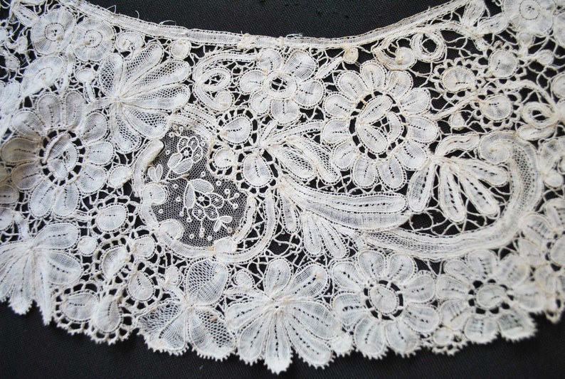 May include: Close-up of a delicate, off-white lace collar. The intricate design features floral and leaf patterns, with a scalloped edge. The lace is finely detailed, showcasing the craftsmanship of the textile.