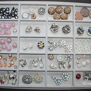 May include: A variety of decorative buttons in a gray divided tray. The buttons are in various shapes, sizes, and colors, including black, pink, gold, silver, and clear. Some buttons have rhinestones or other embellishments.