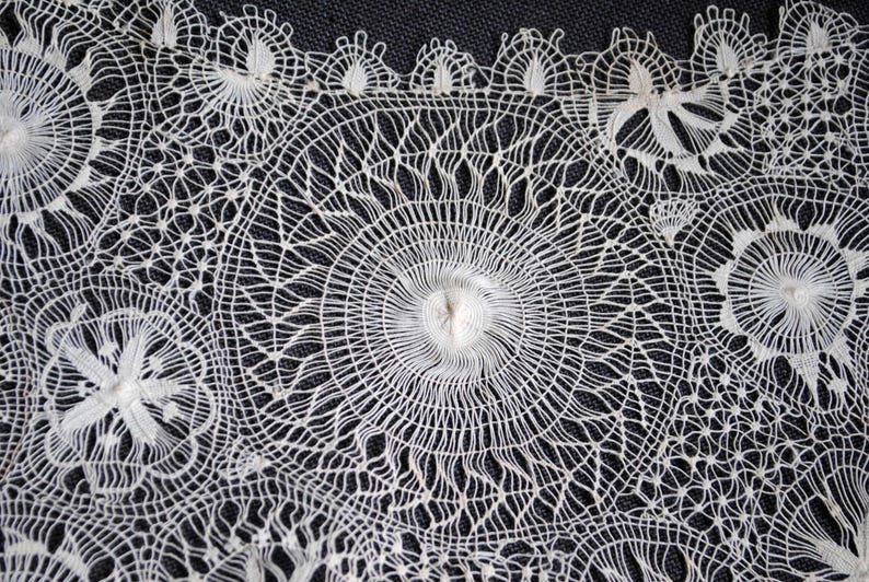 May include: Close-up of white lace with intricate floral and geometric patterns. The lace features circular and star-like designs, with a delicate, open weave. The background is a dark, textured fabric, highlighting the lace's details.