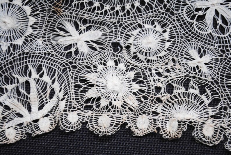 May include: Close-up of a section of white lace with intricate floral and geometric patterns. The lace features circular and fan-shaped designs, creating a delicate and textured appearance. The detailed craftsmanship is evident in the fine, interconnected threads. This textile is suitable for various craft projects.