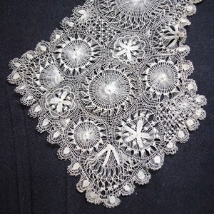 May include: Close-up of a white lace collar with a detailed, circular design. The lace features various floral and geometric patterns, with a scalloped edge. The delicate textile is likely handmade, showcasing intricate needlework.