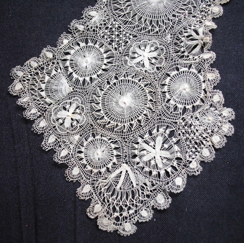 May include: Close-up of a white lace collar with a detailed, circular design. The lace features various floral and geometric patterns, with a scalloped edge. The delicate textile is likely handmade, showcasing intricate needlework.