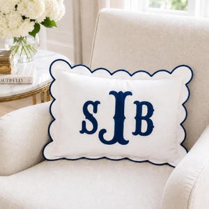 May include: A rectangular white throw pillow featuring a scalloped edge trimmed in navy blue piping. The center of the pillow displays a large, embroidered navy blue monogram with the letters S, J, and B in a decorative serif font style.