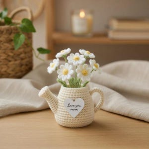 May include: A crocheted watering can vase holds white and yellow daisy flowers. A heart-shaped tag on the vase reads "Love you mom." The vase is beige and sits on a wooden surface. A candle and basket are in the background.