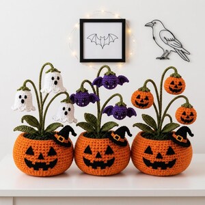 May include: Three crocheted Halloween-themed potted plants in orange pumpkin-shaped pots with black jack-o'-lantern faces and small witch hats. The plants feature white ghosts, purple bats, and orange pumpkins on green stems with leaves.
