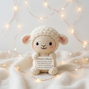 May include: A crocheted white sheep doll with pink accents on the ears and cheeks, holding a sign that reads "Positive Sheep." The sign includes the text "I may be a tiny sheep, but I believe in you. Go do your thing! YOU'RE AWESOME!"