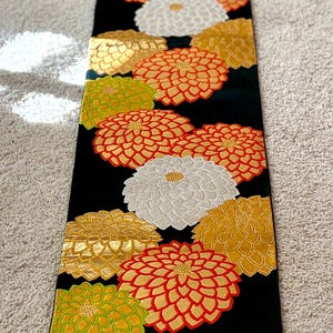 May include: A long, rectangular black fabric runner featuring an embroidered pattern of large, stylized chrysanthemum flowers in vibrant shades of orange, gold, lime green, and white, with a thin horizontal gold metallic thread border near the bottom.
