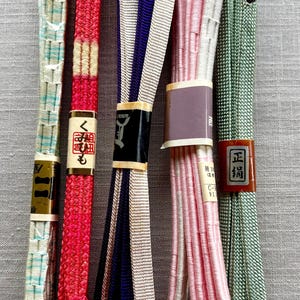 May include: Five long, braided obijime cords for kimono, each featuring distinct colors including light blue, red with cream, purple and beige, light pink, and sage green. Each cord is secured with a paper band, some displaying Japanese text.