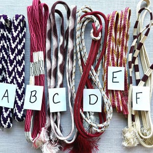 May include: Six long, braided fabric cords labeled A through F. Cord A is purple and white chevron. B is red with white sections. C is wavy brown and white. D is red with white flecks. E is red, gold, and white. F is cream with brown bands.