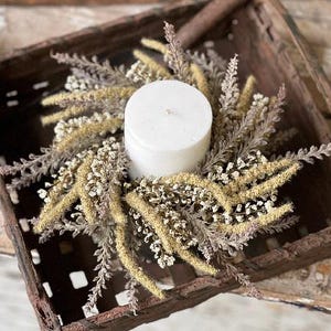 May include: A white pillar candle is centered in a decorative wreath made of dried flowers and foliage. The wreath features a mix of beige, light green, and lavender-colored elements. The candle and wreath are displayed in a rustic, brown wooden basket.