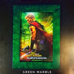 May include: A green marble frame with a movie poster inside. The poster is for the movie "Thor: Ragnarok" and features a character from the movie holding a weapon. The text on the poster reads "Marvel Studios Thor Ragnarok November 3."