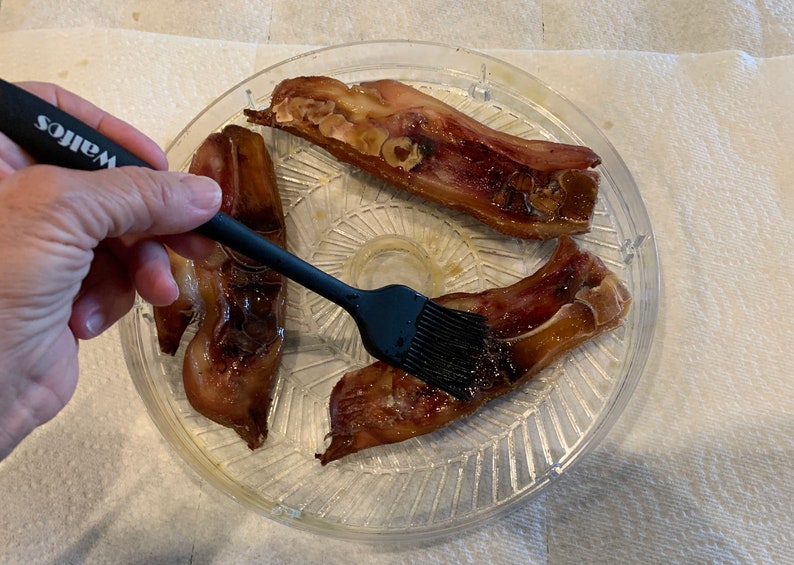 Pode incluir: Uma m&atilde;o segurando um pincel de silicone preto, pincelando uma bandeja de vidro transparente com tr&ecirc;s peda&ccedil;os de carne. A carne &eacute; marrom e brilhante, e a bandeja tem um padr&atilde;o decorativo.