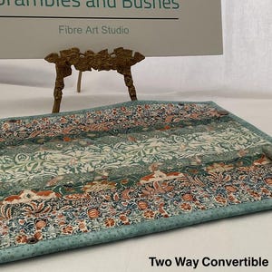 以下が含まれることがあります： ティール、オレンジ、白の色合いの花柄の、2ウェイコンバーチブル生地アイテム。 ブロンズ色のボタンで留められ、ティール色の縁取りがあります。「Two Way Convertible」の文字が見えます。