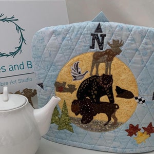 May include: A blue quilted tea cosy with a compass design featuring a bear, moose, and birds. The cosy has a yellow centre with a brown border and a white compass needle pointing north.