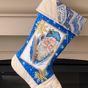 以下が含まれることがあります： 金色の雪の結晶のデザインとサンタクロースのクロスステッチの画像が施された青と白のクリスマスストッキング。暖炉のマントルピースに吊るされています。