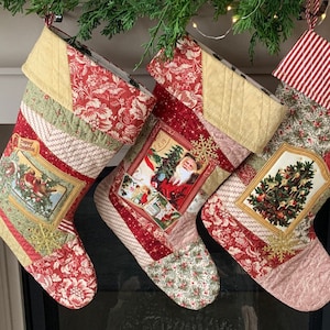 May include: Three Christmas stockings made of patchwork fabric with different holiday scenes. The stockings are red, green and cream with gold accents. The stockings are hanging from a garland of greenery.