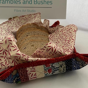 May include: A red and white fabric bread basket with a floral pattern. The basket is filled with slices of brown bread. The basket is labelled "Bramble and Bushes Fibre Art Studio".