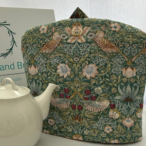 以下が含まれることがあります： 鳥とイチゴが描かれた花柄の緑色のティーコージー。ティーコージーは布製で、伝統的なデザインです。