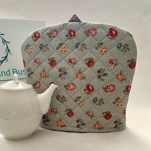 May include: A quilted teapot cosy with a floral pattern in shades of red, pink, yellow, and blue on a light grey background. The cosy has a diamond quilted pattern and white polka dots.