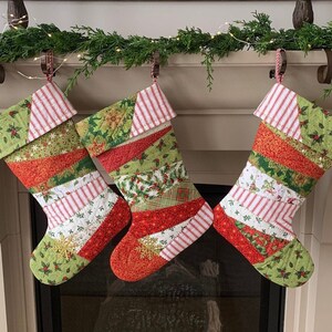以下が含まれることがあります： 白い暖炉の上に、緑の葉と電球のガーランドが飾られた、赤と緑の継ぎ合わせのクリスマスストッキングが3つ吊り下げられています。