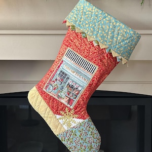 以下が含まれることがあります： 暖炉のマントルピースに、赤と青のキルティング模様のクリスマスストッキングが吊るされています。ストッキングには、おもちゃ屋さんの店の小さな刺繍の絵が描かれており、「The Toy Game」という文字が書かれています。