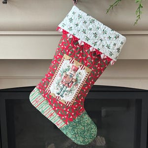 May include: A quilted Christmas stocking featuring a red fabric with a green holly and white berry print. The cuff is white with a small holly pattern. A central rectangular patch displays a nutcracker figure surrounded by evergreen trees.