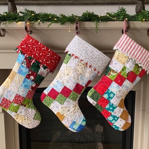 以下が含まれることがあります： 白い暖炉の上に、クリスマスの靴下3つが吊るされています。それぞれの靴下は、赤、緑、青、白、金など、さまざまな色と模様のパッチワーク生地で作られています。靴下は、白いレースのトリムと赤と白のストライプのリボンで飾られています。