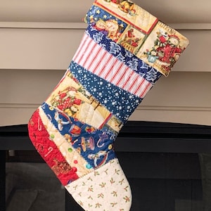 以下が含まれることがあります： パッチワークデザインのキルティングクリスマスストッキング。サンタクロース、星、ストライプ、お祝いのモチーフなど、お祝いのパターンが施されたさまざまな生地が特徴です。つま先はクリーム色でヒイラギ柄で、赤と白のストライプのコードから吊り下げられています。