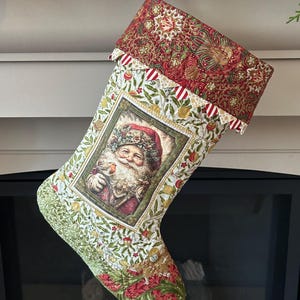 以下が含まれることがあります： サンタクロースの画像が特徴のクリスマスストッキング。赤と緑の花柄模様。カフには赤と金のペイズリー柄。赤と白のストライプのリボンが付いた黒い金属製のフックから吊り下げられています。